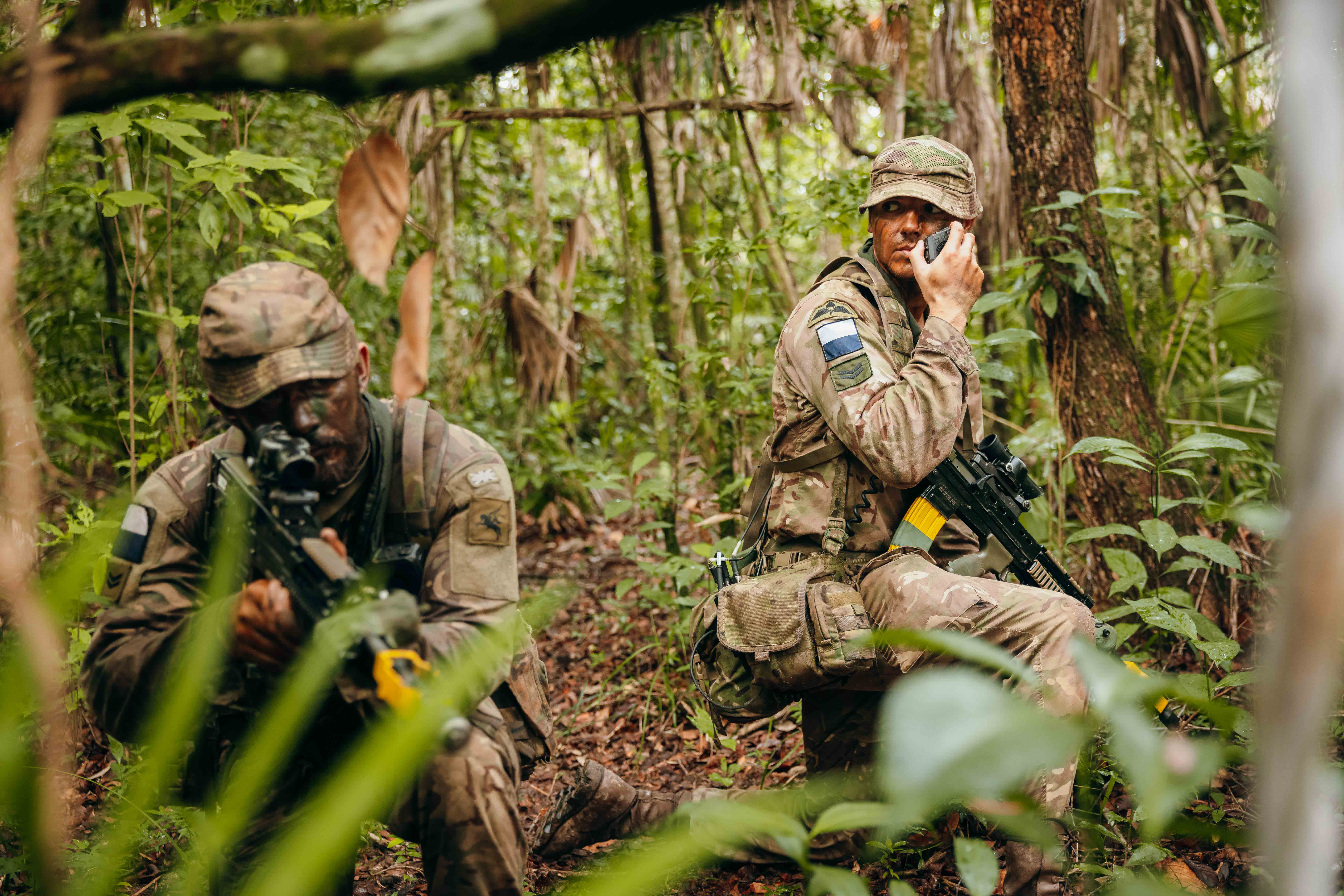 In a jungle environment, two British Army soldiers wearing camouflage uniforms, are kneeling on one knee. The soldier on the left has a rifle, and is looking down the scope. The soldier on the right is talking into a radio communication device.  