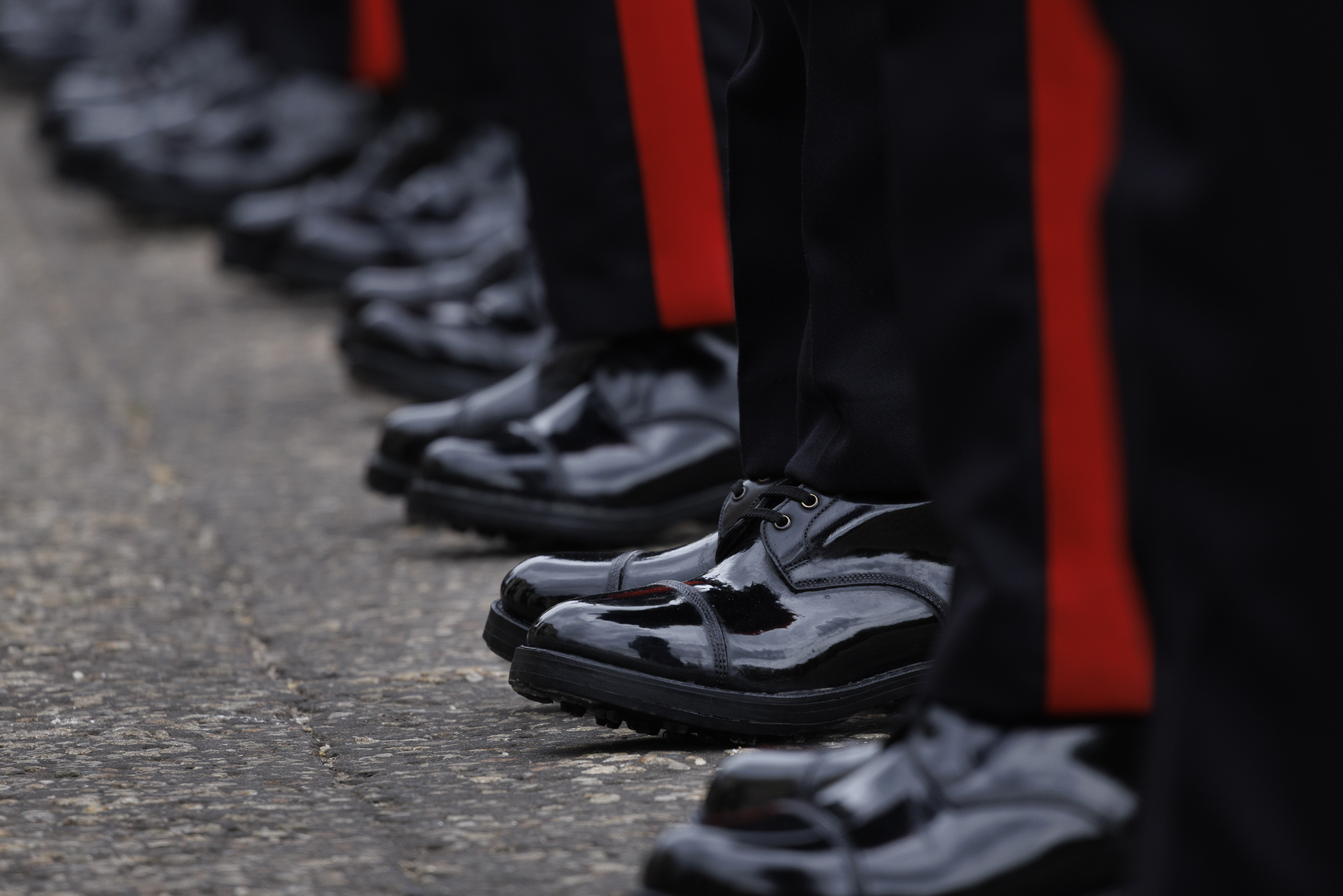 A row of black, polished dress shoes with red-striped trousers, worn by individuals.