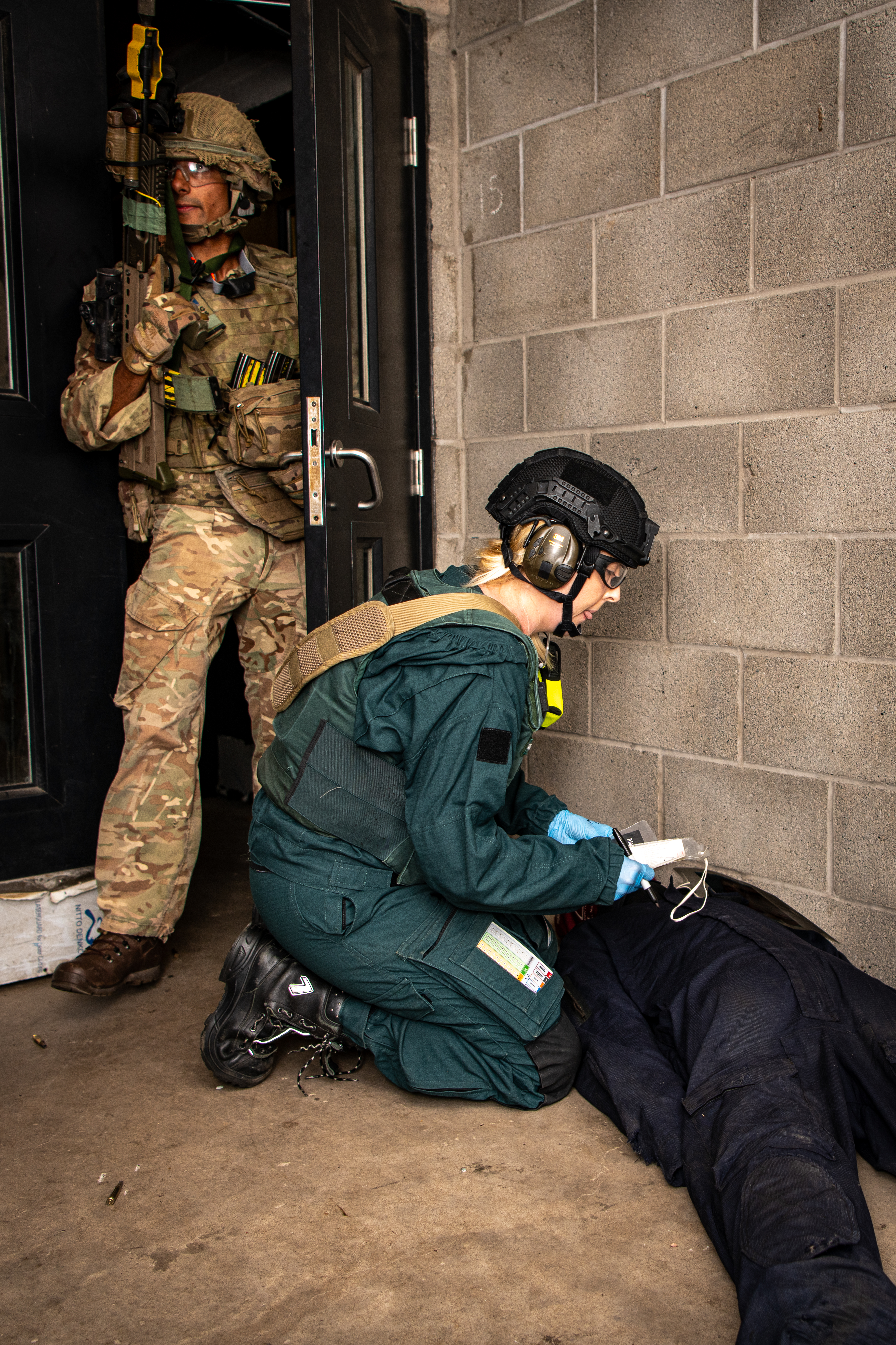 An emergency responder in tactical gear tends to a person lying on the ground, while another uniformed person stands in a doorway holding a firearm. The setting appears to be a concrete-walled indoor area.