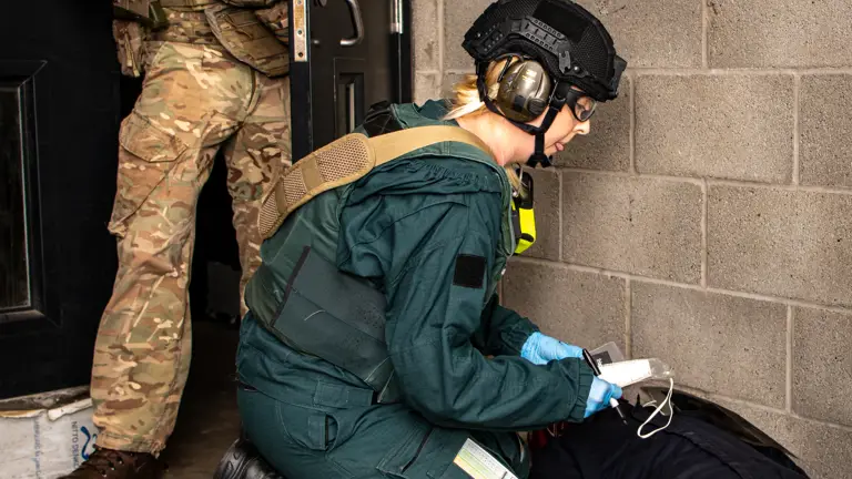 An emergency responder in tactical gear tends to a person lying on the ground, while another uniformed person stands in a doorway holding a firearm. The setting appears to be a concrete-walled indoor area.
