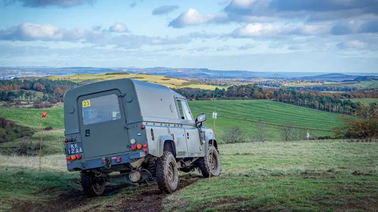 Off-road vehicle on a grassy hilltop, surrounded by rolling green fields under a blue sky with scattered clouds, evoking a sense of adventure.