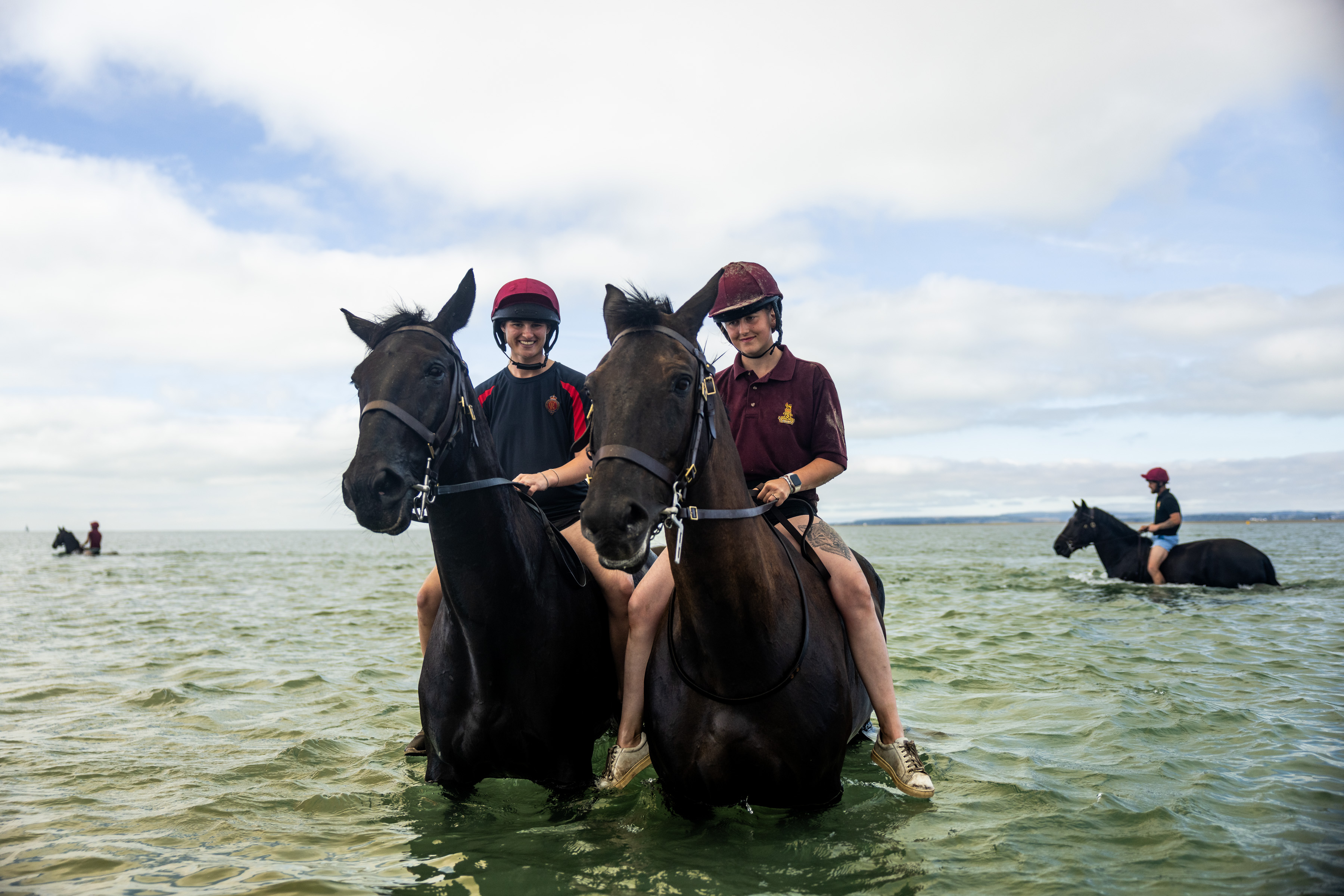 Three people are on horseback in a shallow body of water, with two in the foreground and one in the background. The horses are dark-colored, and all riders are wearing helmets. The sky is partly cloudy.