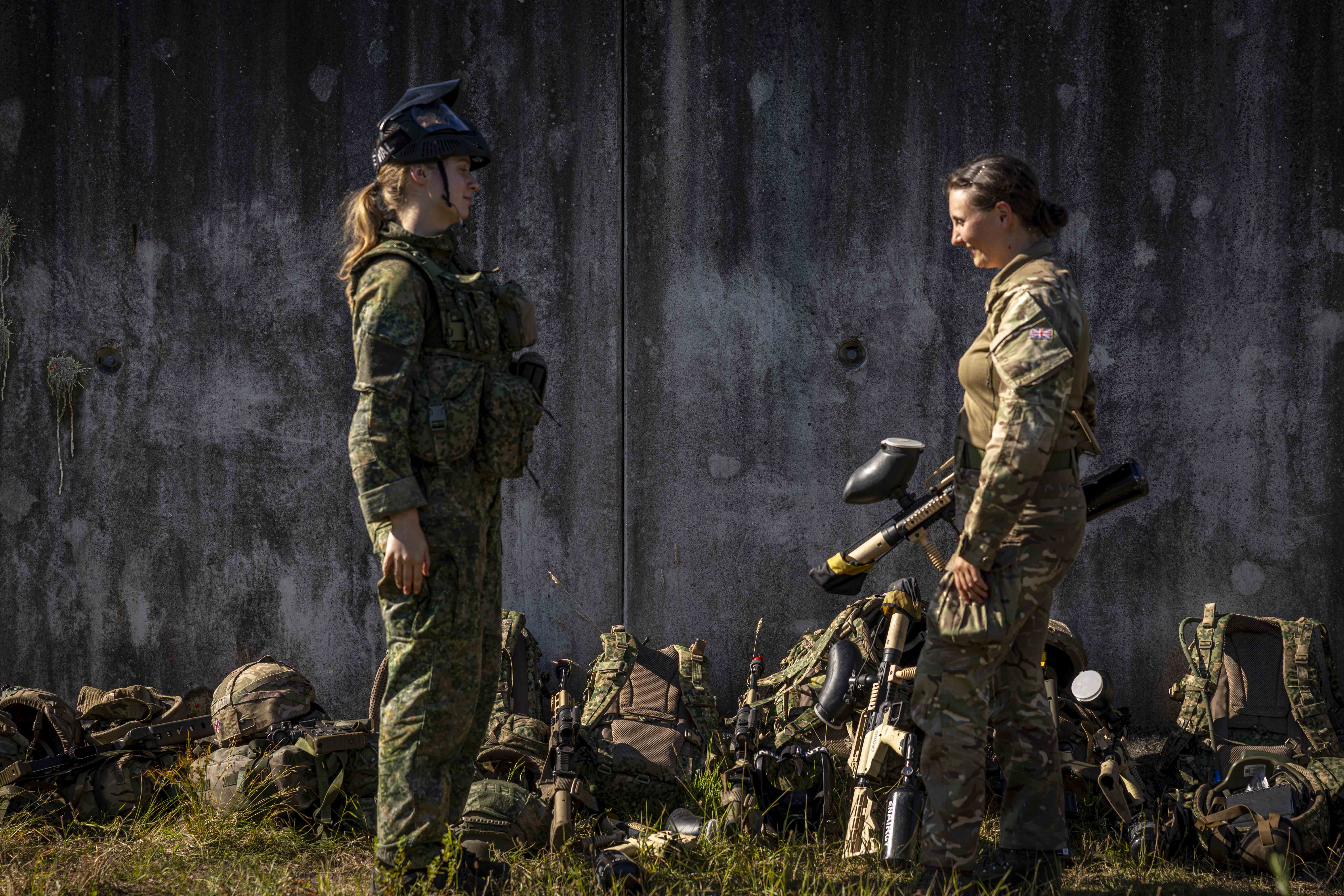 Two individuals in military gear stand facing each other in front of a weathered wall. Several military backpacks and equipment are on the ground nearby. Both are wearing camouflage uniforms and hold paintball markers. Grass is visible in the foreground.