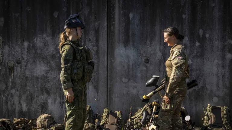 Two individuals in military gear stand facing each other in front of a weathered wall. Several military backpacks and equipment are on the ground nearby. Both are wearing camouflage uniforms and hold paintball markers. Grass is visible in the foreground.