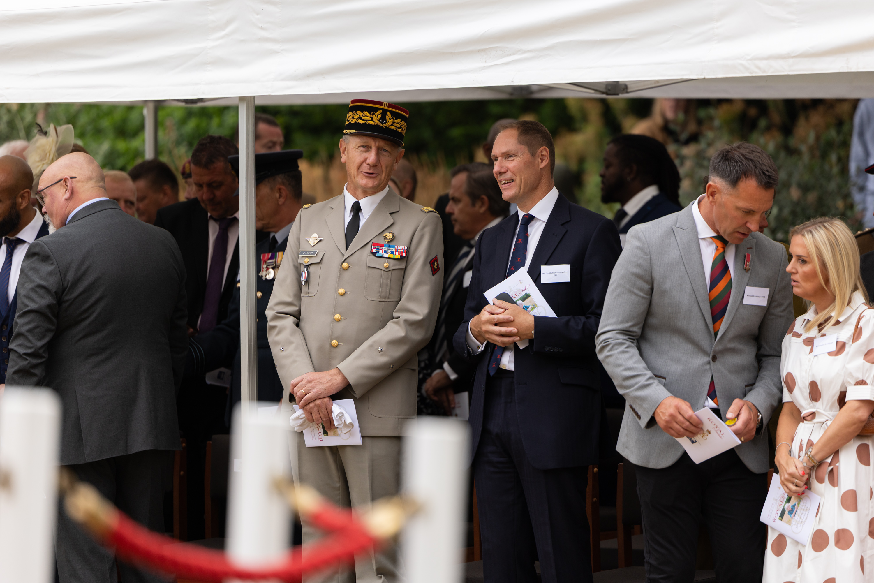 A group of people are gathered under a canopy, engaged in conversation. One person is wearing a military uniform.