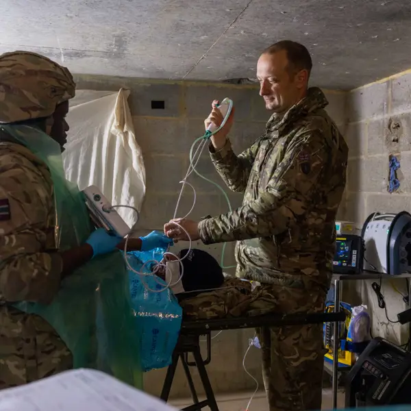 Two soldiers in camouflage uniforms conduct a medical procedure in a concrete-walled room.