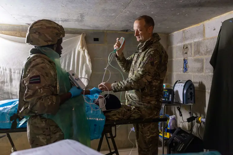 Two soldiers in camouflage uniforms conduct a medical procedure in a concrete-walled room.