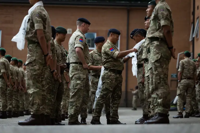 Soldiers are standing in two rows, waiting to receive a white scarf tied around their necks from British Army officers.