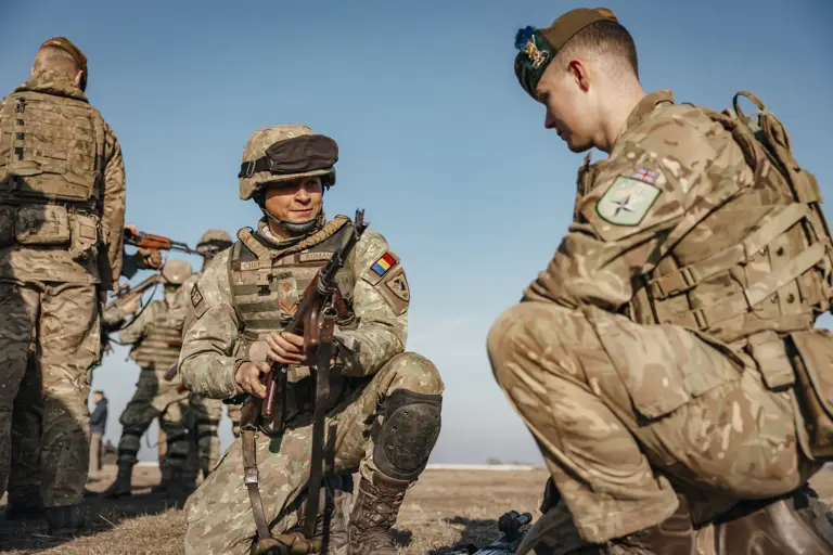 A Romanian soldier and British soldier showcased each other’s weapons including rifles on the ground outside. Both soldiers are wearing camouflage uniform and headgear in their retrospective country uniforms.
