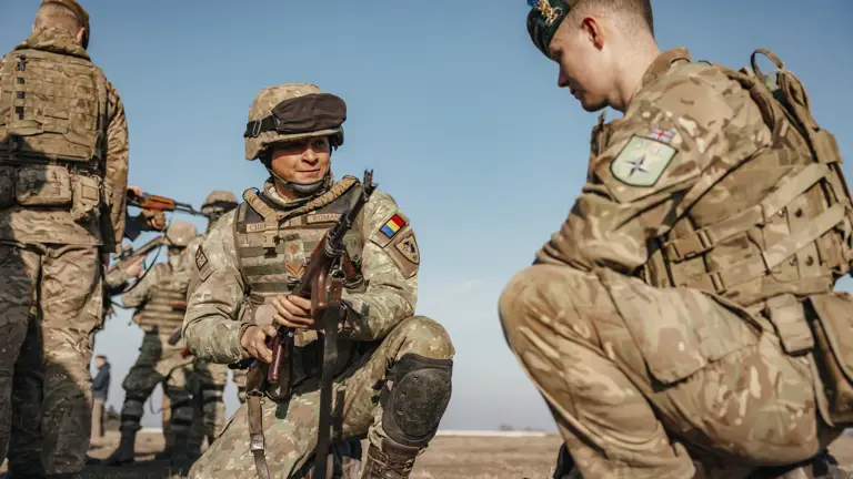 A Romanian soldier and British soldier showcased each other’s weapons including rifles on the ground outside. Both soldiers are wearing camouflage uniform and headgear in their retrospective country uniforms.