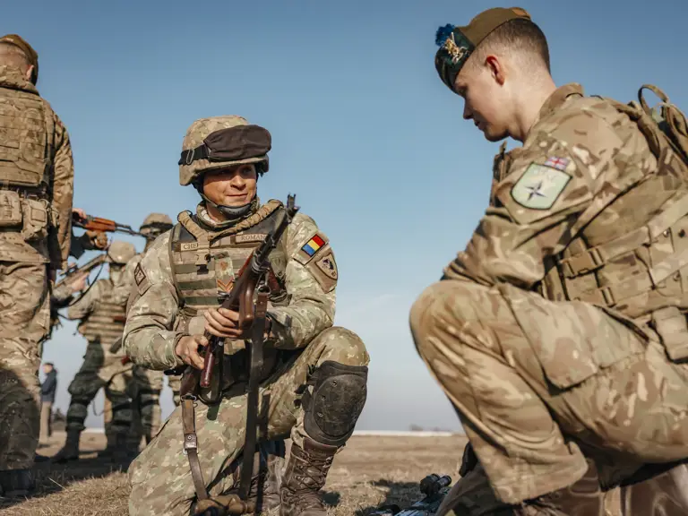 A Romanian soldier and British soldier showcased each other’s weapons including rifles on the ground outside. Both soldiers are wearing camouflage uniform and headgear in their retrospective country uniforms.