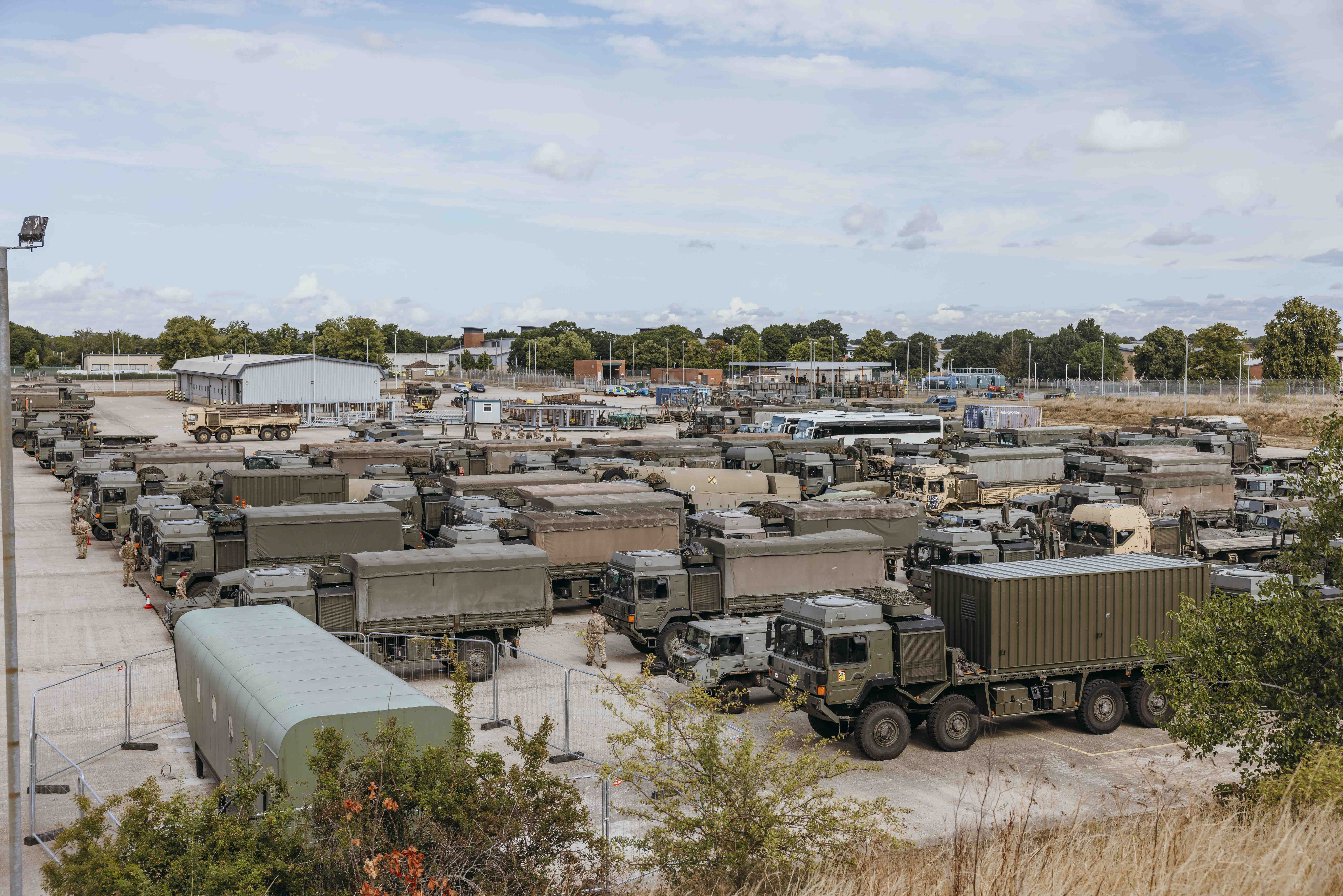 Green military trucks are parked in rows. Soldiers are walking between the trucks.