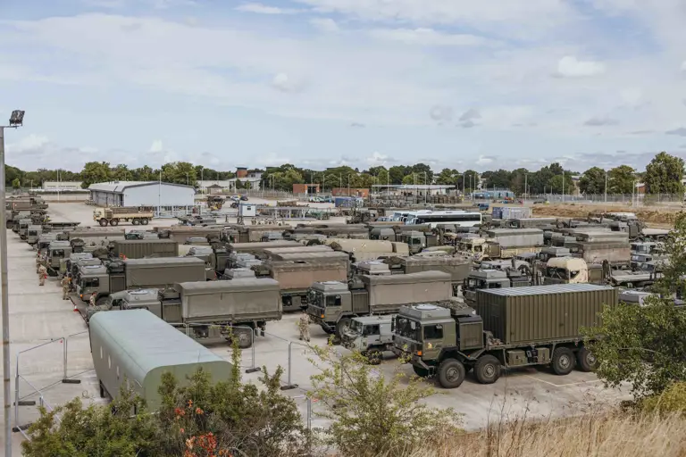 Green military trucks are parked in rows. Soldiers are walking between the trucks.