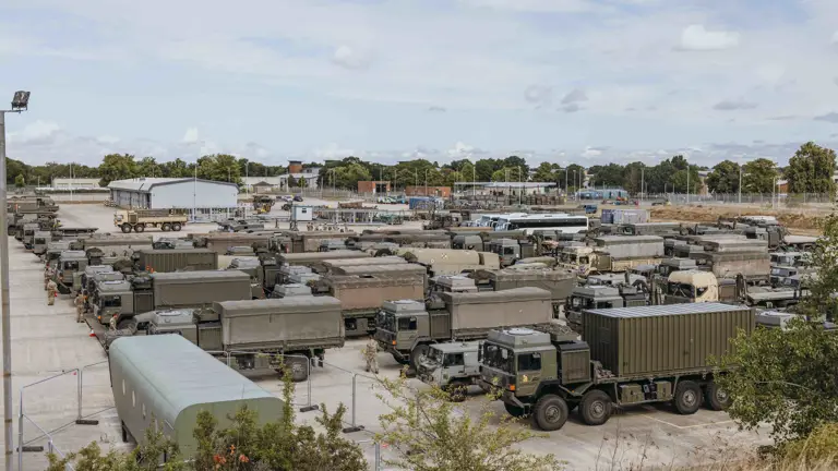 Green military trucks are parked in rows. Soldiers are walking between the trucks.
