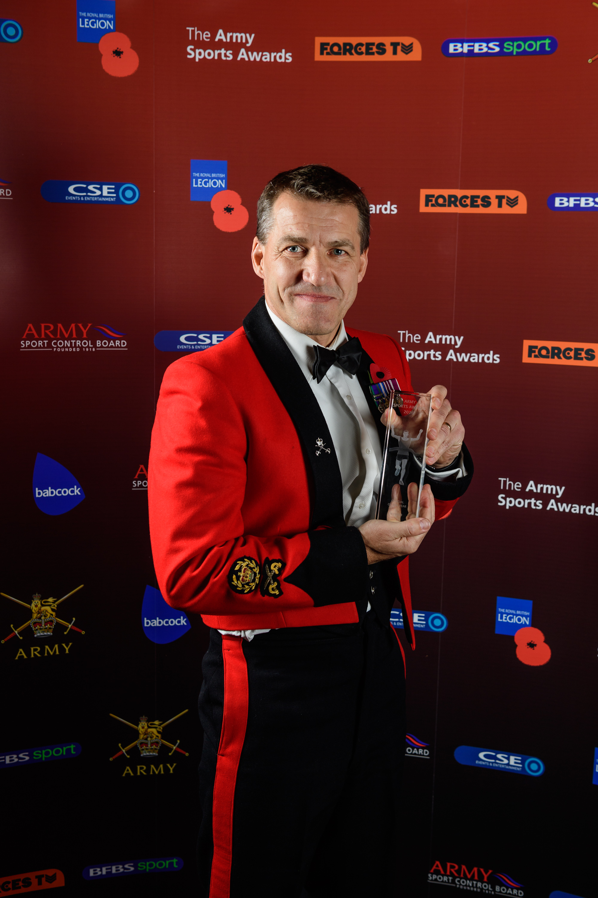 This image shows a person in a formal red and black uniform holding a trophy against a backdrop with logos.