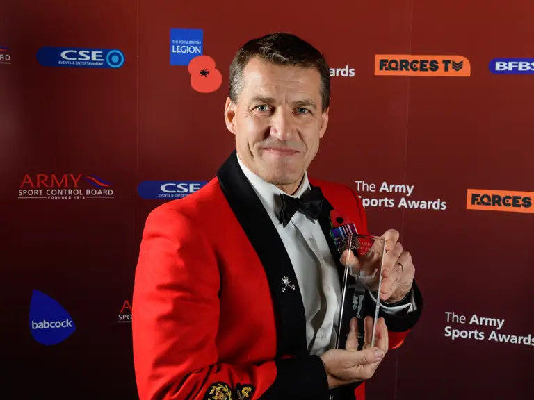 This image shows a person in a formal red and black uniform holding a trophy against a backdrop with logos.