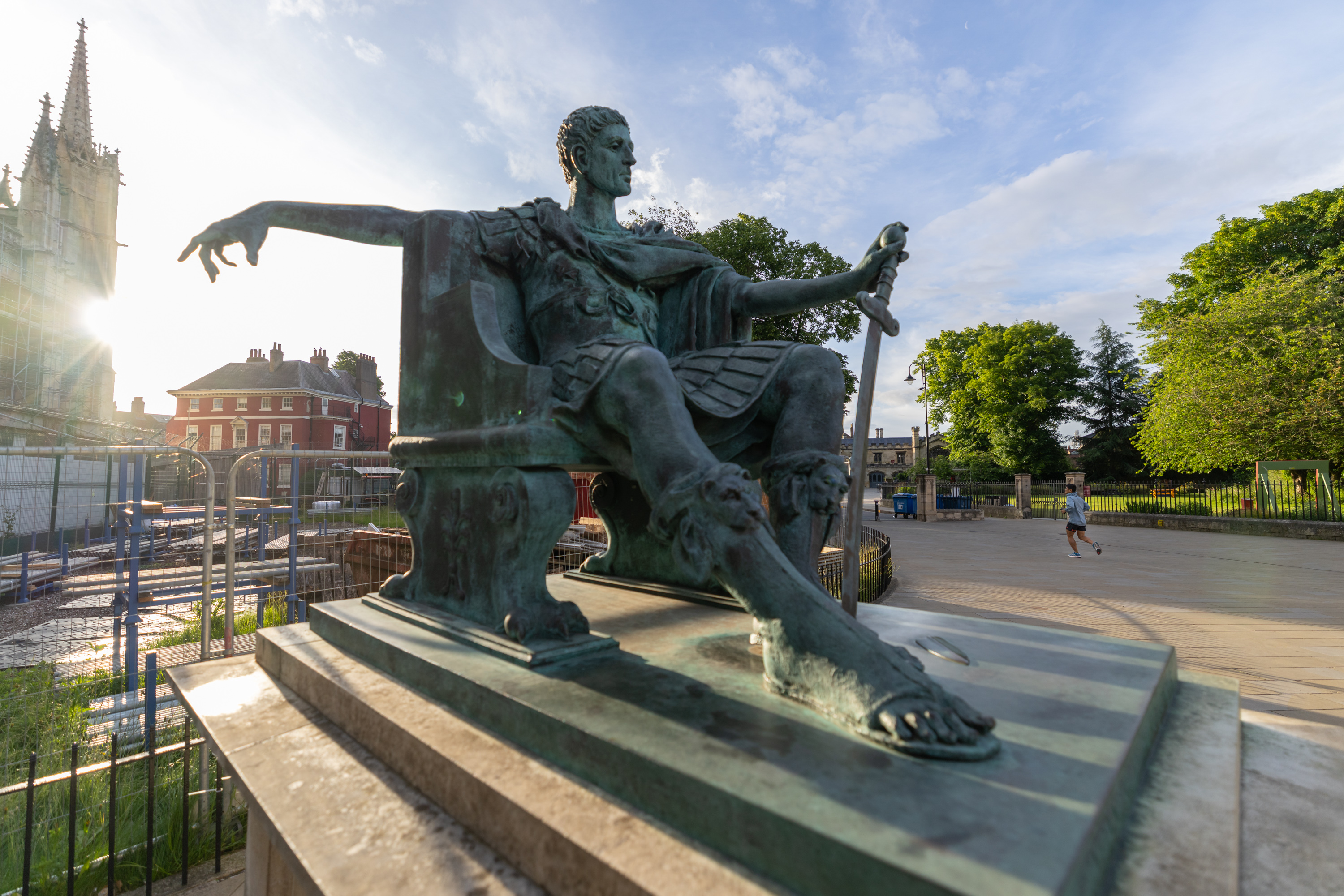 A large bronze statue of a seated ancient figure with a draped garment and sandals, holding a downward-pointing sword. The figure overlooks a paved area with York Minster in the background. A few pedestrians are visible nearby, surrounded by trees and buildings.