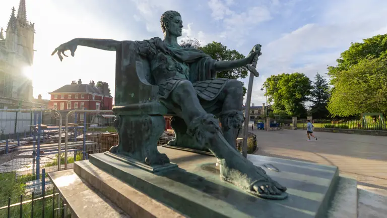 A large bronze statue of a seated ancient figure with a draped garment and sandals, holding a downward-pointing sword. The figure overlooks a paved area with York Minster in the background. A few pedestrians are visible nearby, surrounded by trees and buildings.
