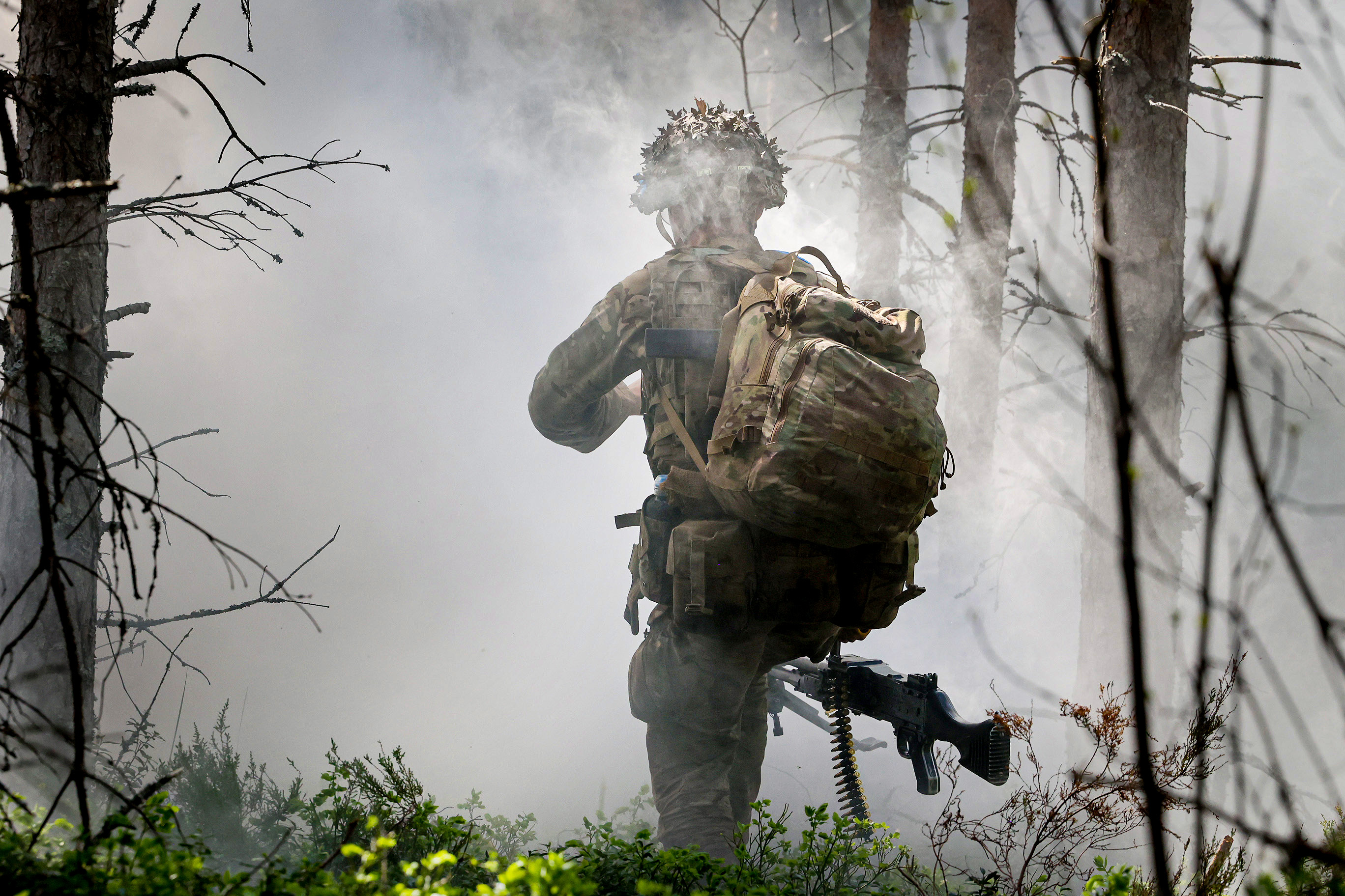 A person in military gear and a large backpack moves through a smoky forest area. They carry a rifle and wear a helmet.