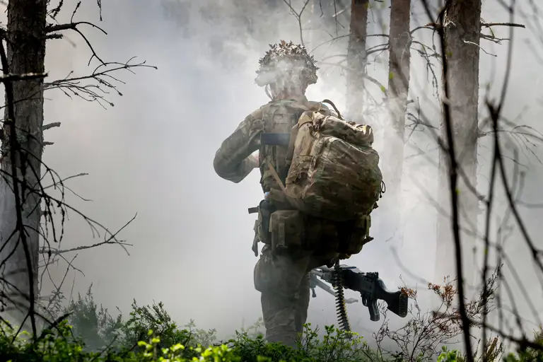 A person in military gear and a large backpack moves through a smoky forest area. They carry a rifle and wear a helmet.