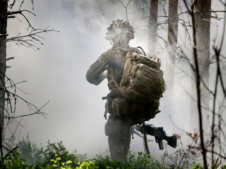 A person in military gear and a large backpack moves through a smoky forest area. They carry a rifle and wear a helmet.