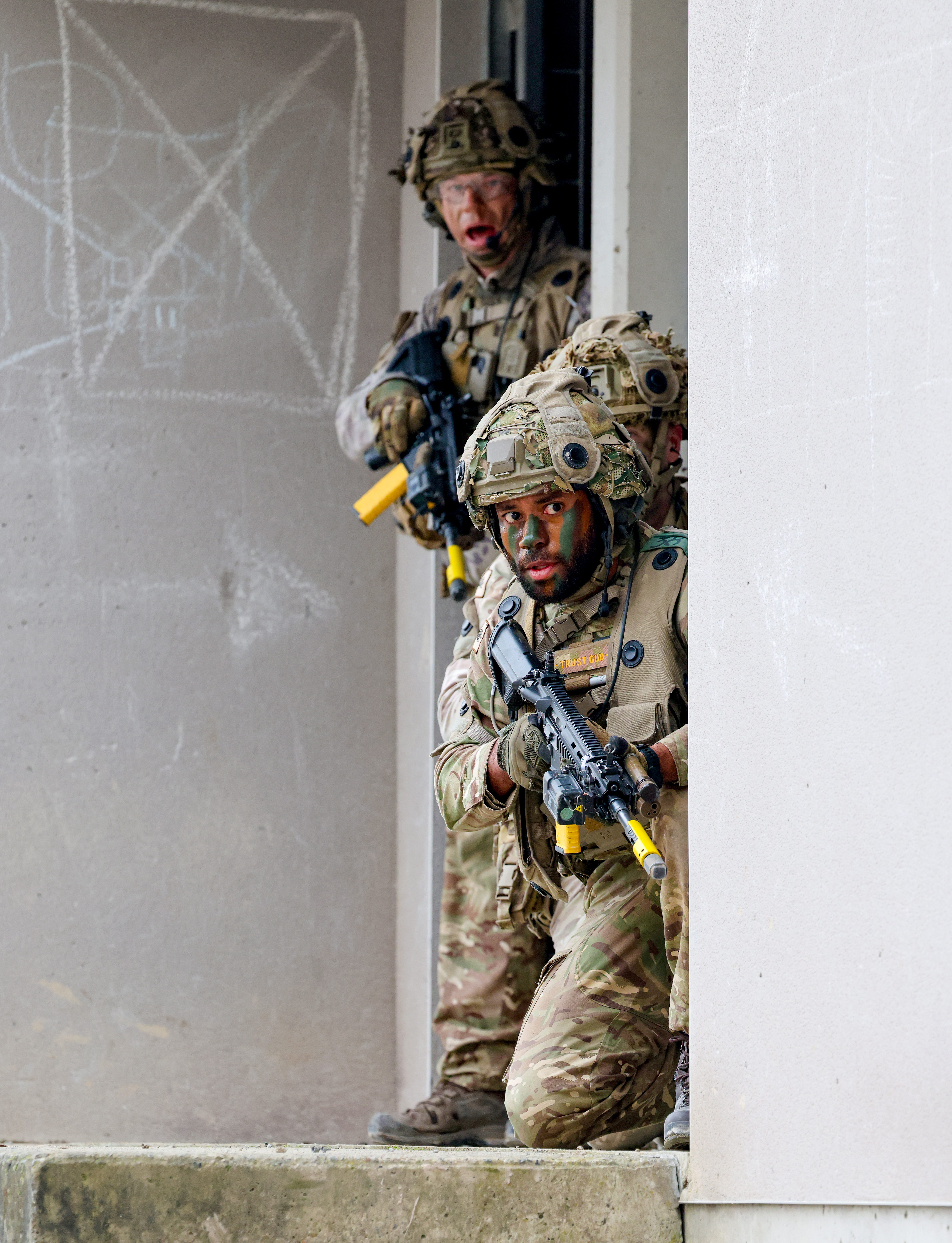 Two soldiers in camouflage gear and tactical helmets are positioned at the corner of a building, holding rifles.