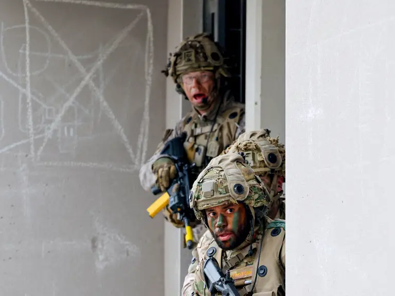 Two soldiers in camouflage gear and tactical helmets are positioned at the corner of a building, holding rifles.