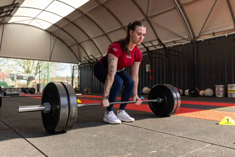 A woman in athletic wear, focused, prepares to lift a heavy barbell in a gym with a high ceiling. The space has red and black flooring, creating a determined atmosphere.