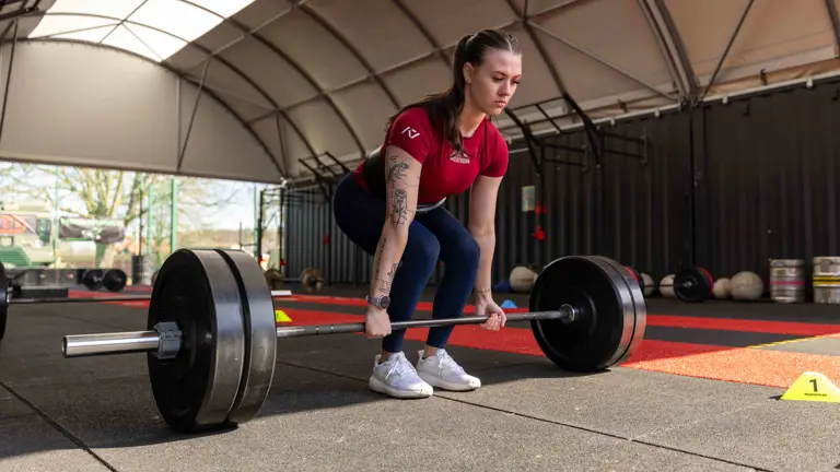 A woman in athletic wear, focused, prepares to lift a heavy barbell in a gym with a high ceiling. The space has red and black flooring, creating a determined atmosphere.
