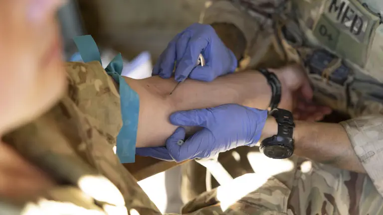 An individual wearing camouflage military attire is having blood drawn from an arm by another person in similar uniform, with blue gloves. The setting appears to be a medical or field environment.