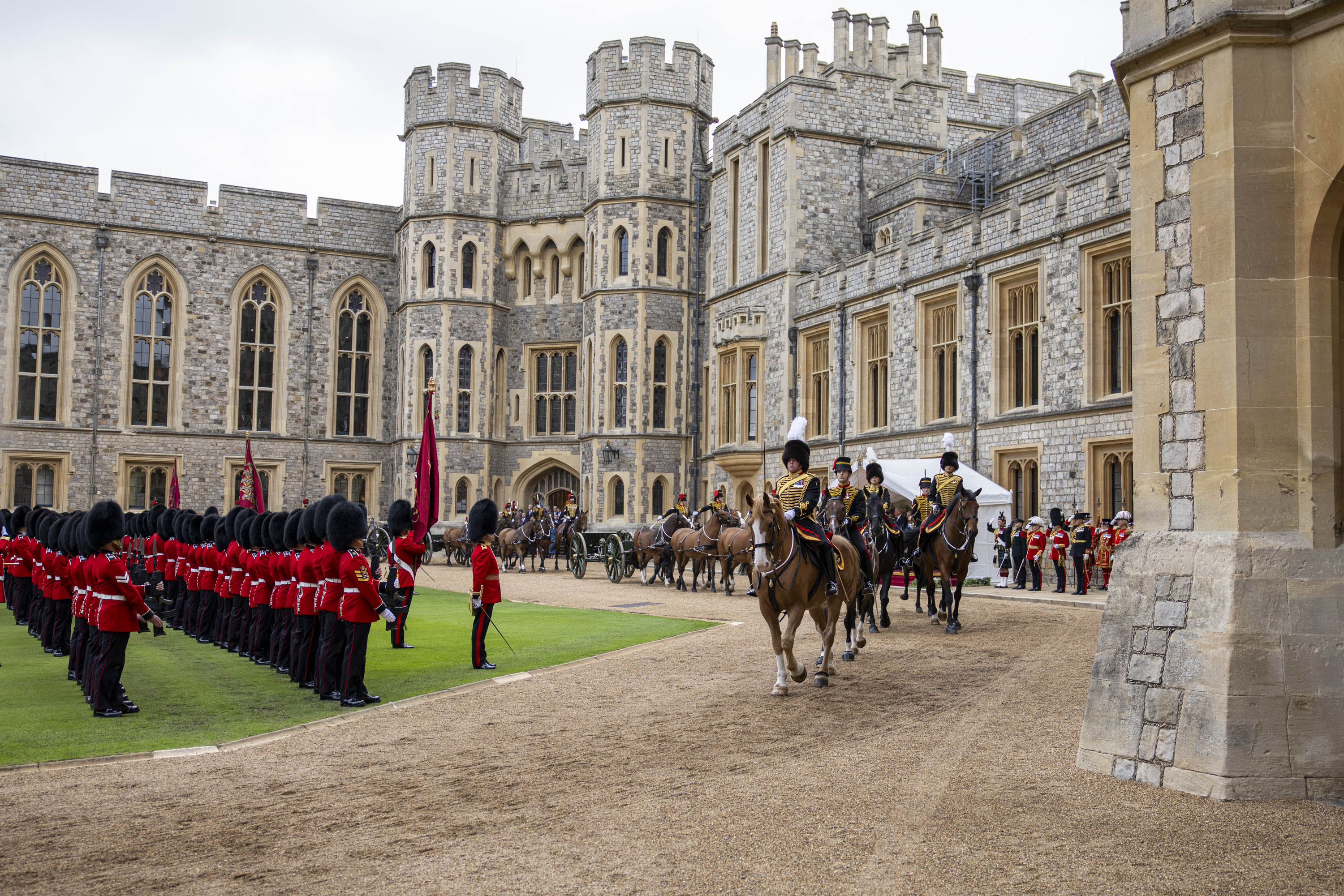 Mounted guards in ceremonial uniforms ride horses past a formation of soldiers in red coats and bearskin hats.