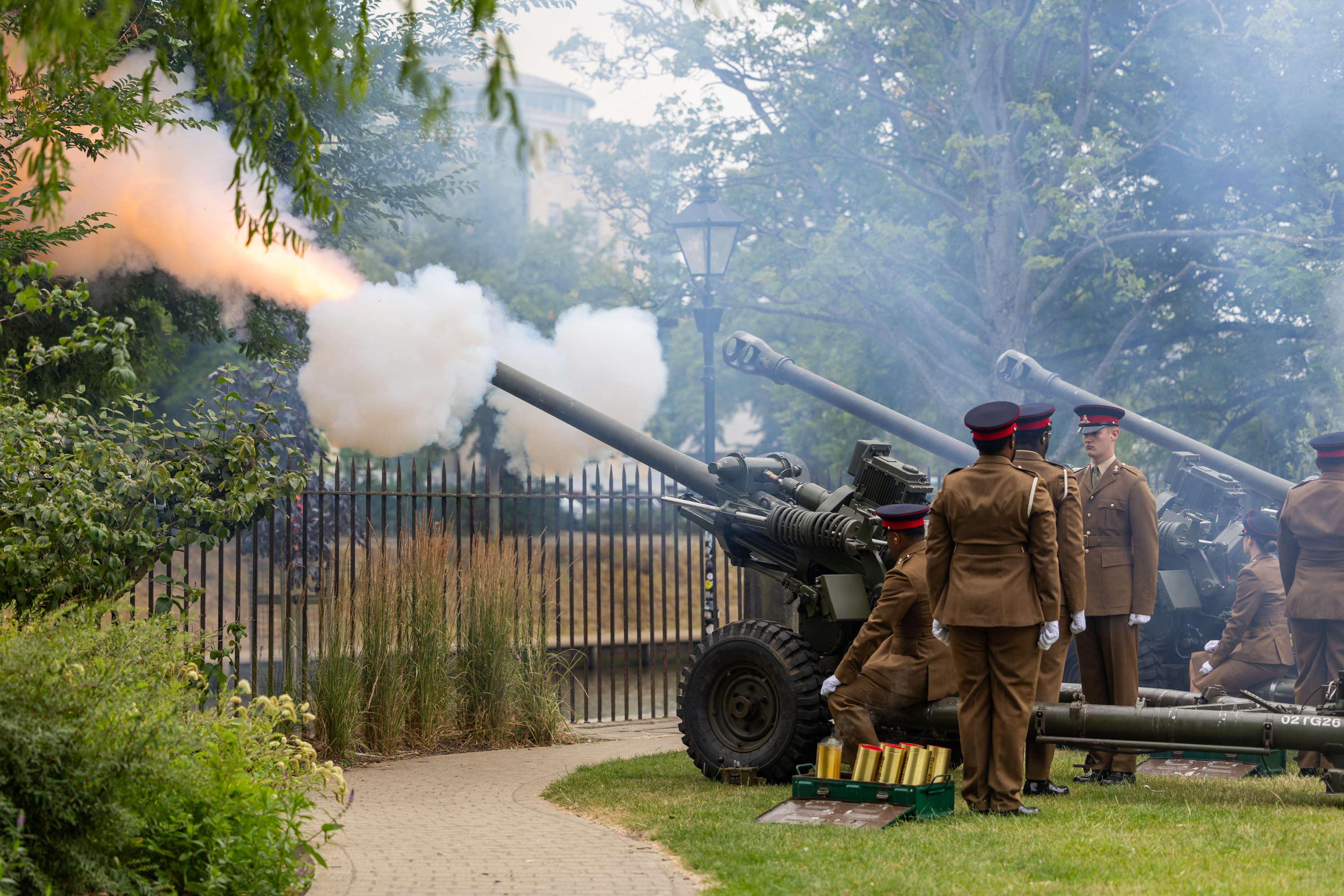 A group of people in brown military uniforms are operating three cannons in a park setting. One cannon is firing.