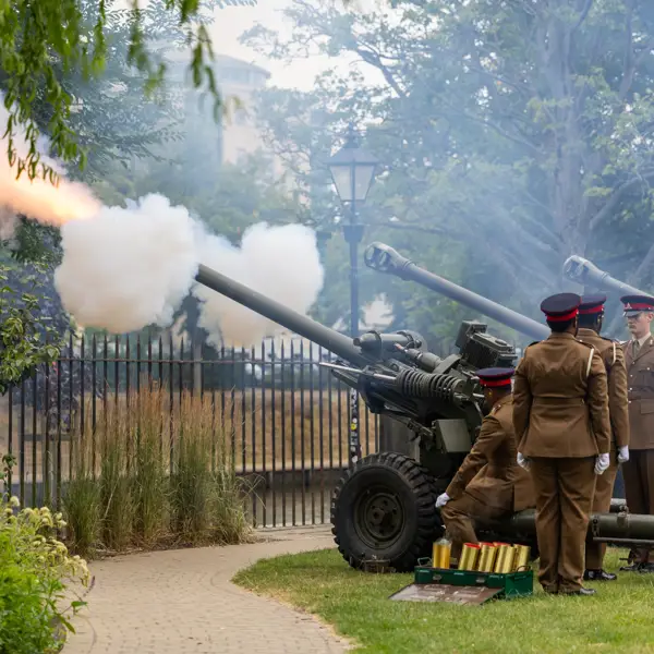 A group of people in brown military uniforms are operating three cannons in a park setting. One cannon is firing.