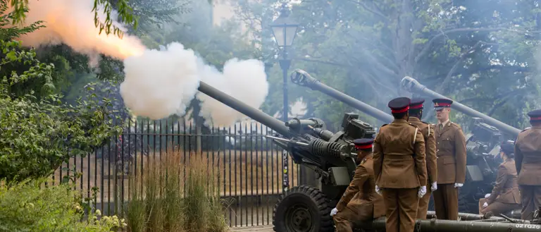 A group of people in brown military uniforms are operating three cannons in a park setting. One cannon is firing.