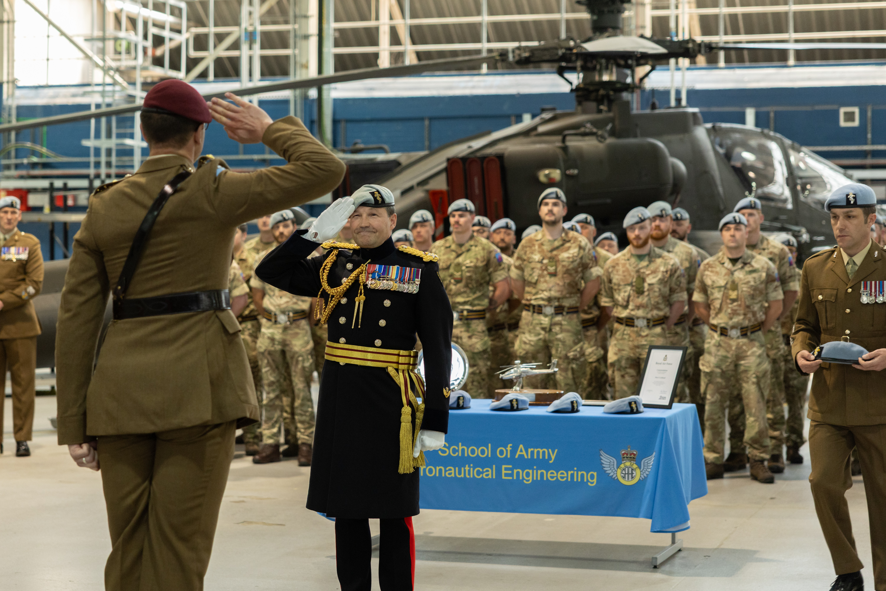 Two soldiers, one in brown ceremonial uniform and one in black ceremonial uniform, salute each other in front of a green military helicopter.