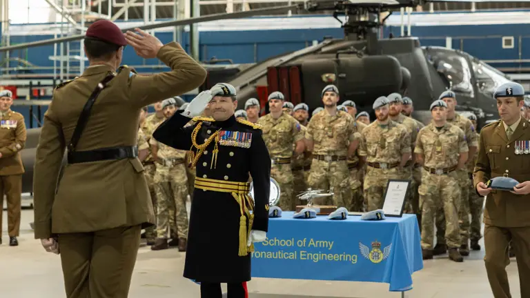 Two soldiers, one in brown ceremonial uniform and one in black ceremonial uniform, salute each other in front of a green military helicopter.