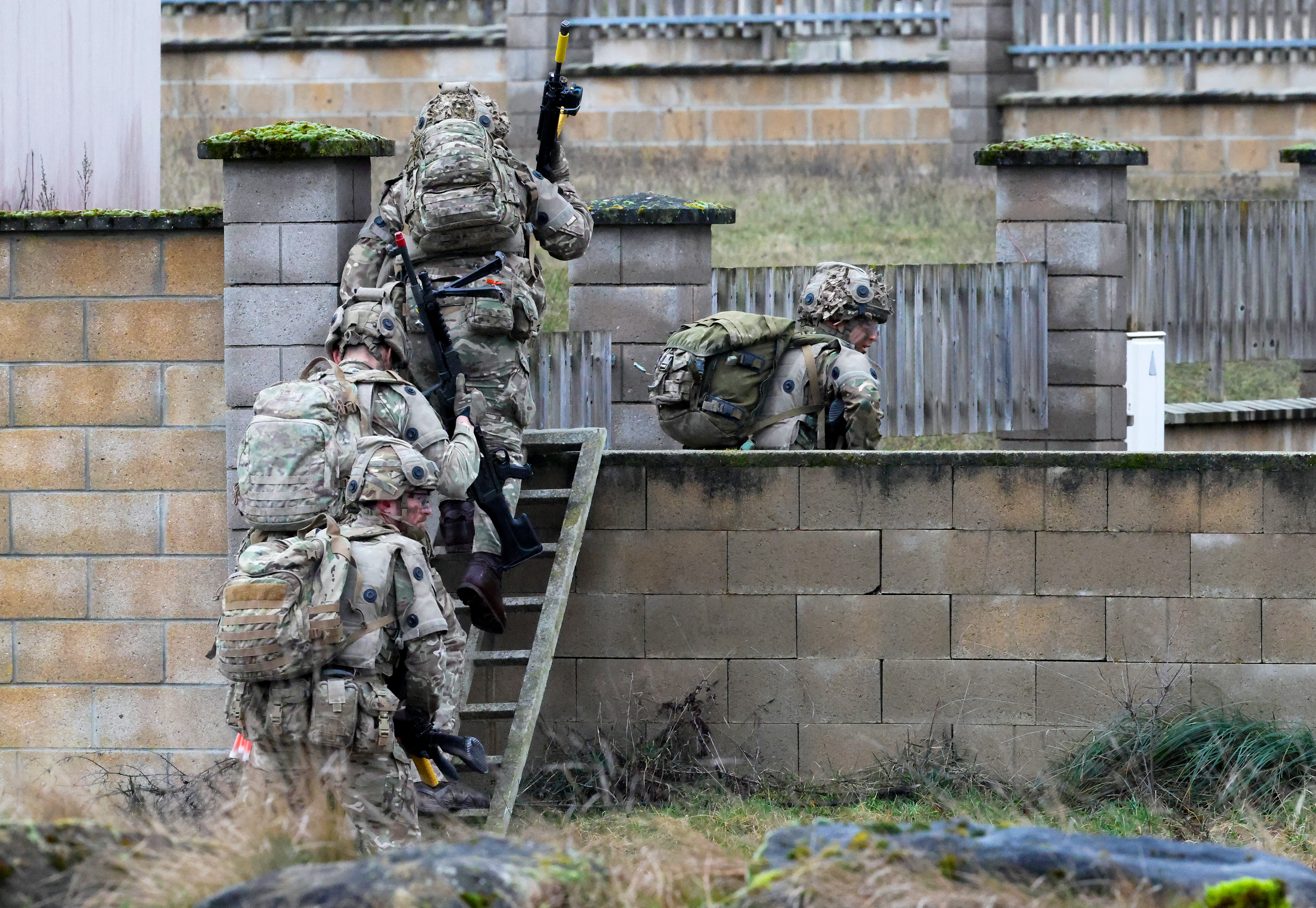A group of soldiers in camouflage uniforms are carrying equipment while climbing over a stone wall.
