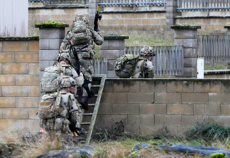 A group of soldiers in camouflage uniforms are carrying equipment while climbing over a stone wall.