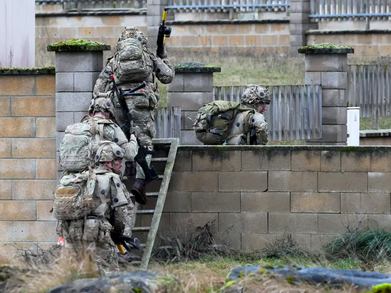 A group of soldiers in camouflage uniforms are carrying equipment while climbing over a stone wall.