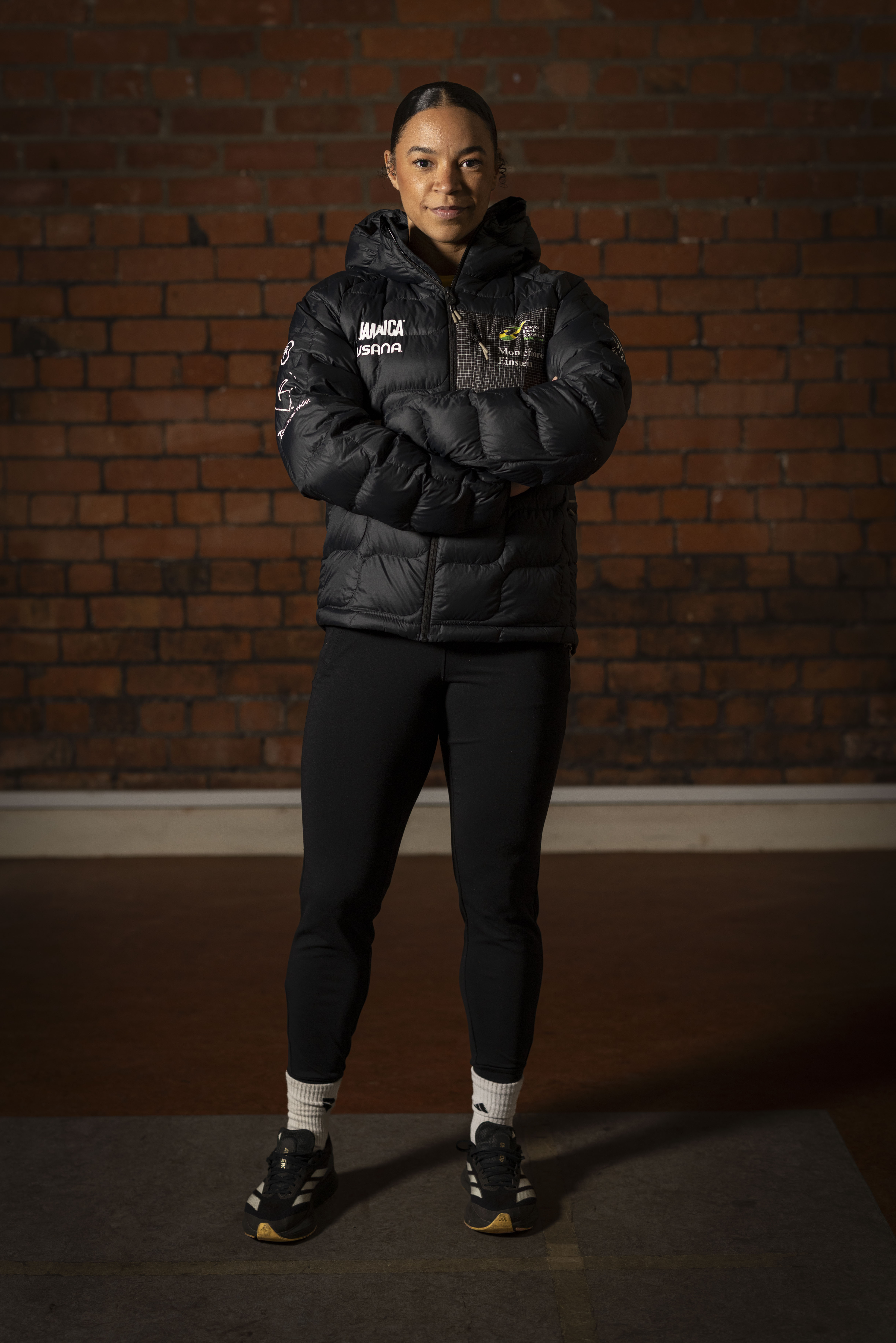 Person standing with arms crossed wearing a black puffer jacket with the words 'Jamaica bobsled', black leggings, and sneakers against a brick wall background.