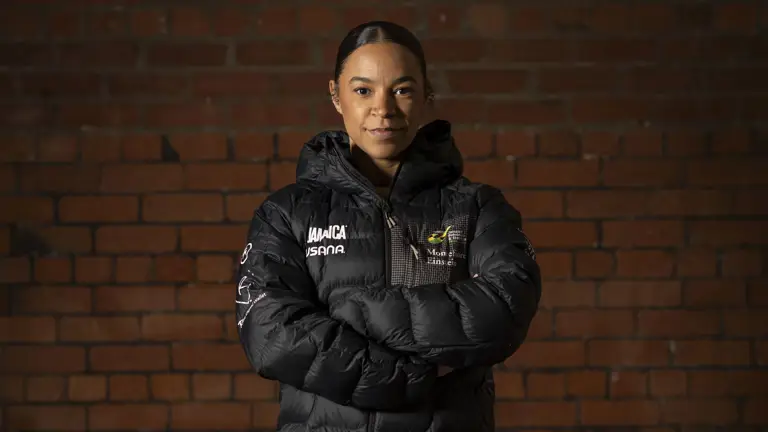 Person standing with arms crossed wearing a black puffer jacket with the words 'Jamaica bobsled', black leggings, and sneakers against a brick wall background.