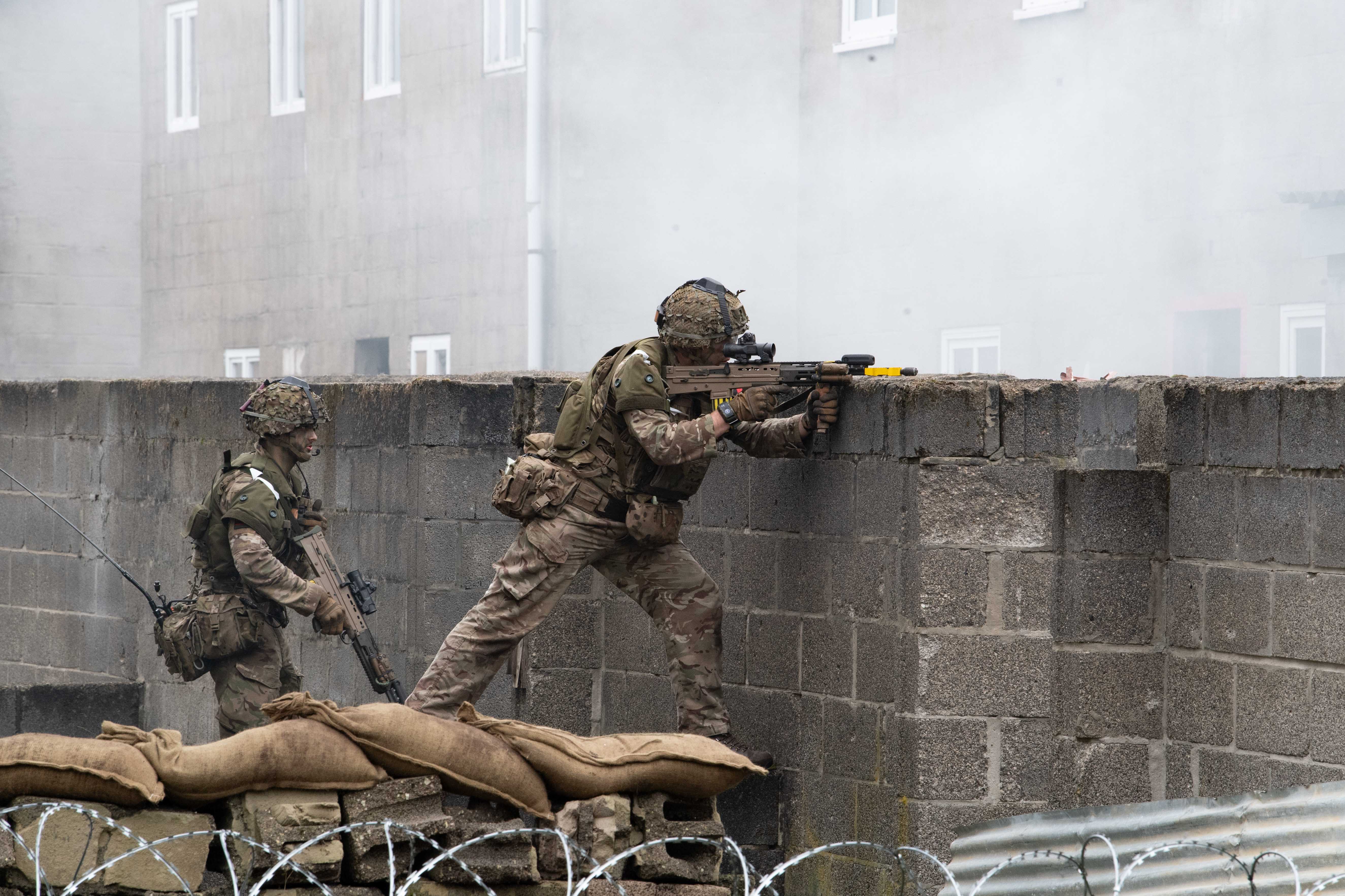 Two soldiers in camouflage gear aim rifles over a brick wall amidst smoke. Sandbags and barbed wire create a tense, combat-like atmosphere.