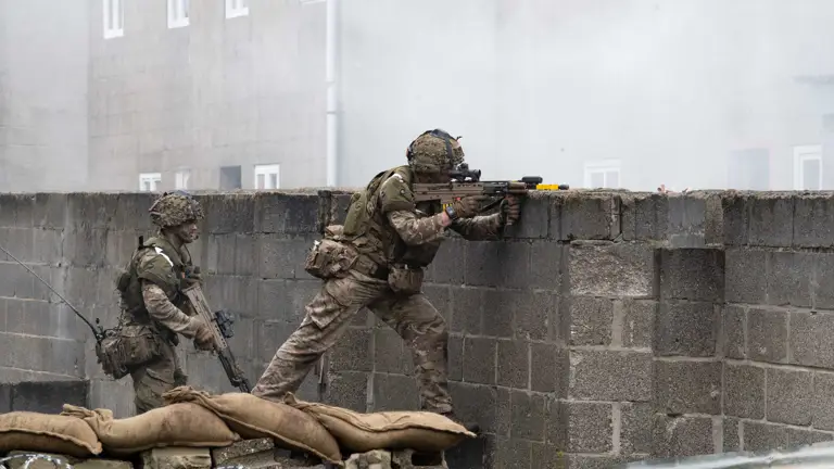 Two soldiers in camouflage gear aim rifles over a brick wall amidst smoke. Sandbags and barbed wire create a tense, combat-like atmosphere.