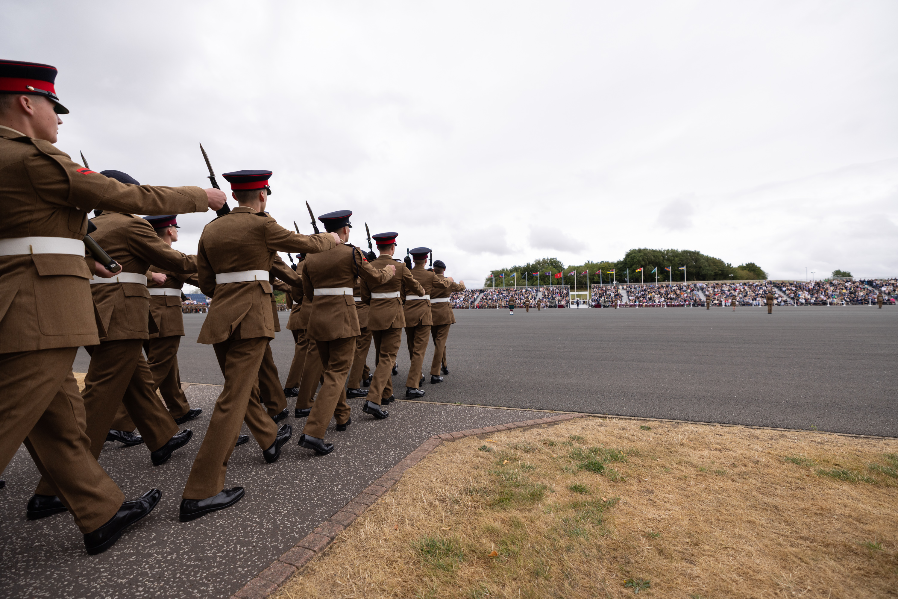 A group of uniformed individuals march in formation on a paved area, holding rifles. A large crowd is seated in the background, with numerous flags visible.