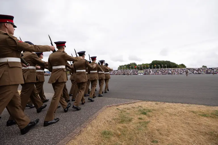 A group of uniformed individuals march in formation on a paved area, holding rifles. A large crowd is seated in the background, with numerous flags visible.