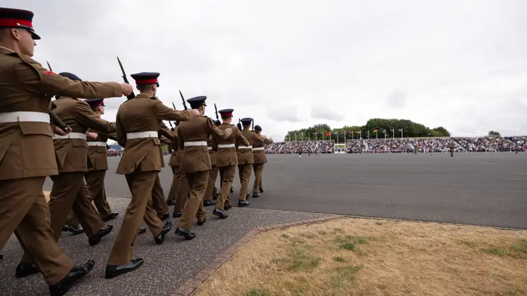 A group of uniformed individuals march in formation on a paved area, holding rifles. A large crowd is seated in the background, with numerous flags visible.
