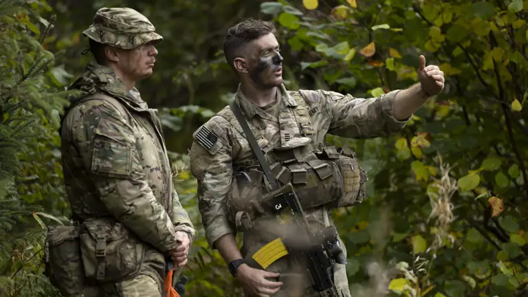 Two individuals in military camouflage uniforms are standing in a forest. One is pointing towards something in the distance. Both have tactical gear, and one has face paint. The scene is surrounded by green foliage.