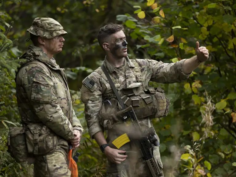 Two individuals in military camouflage uniforms are standing in a forest. One is pointing towards something in the distance. Both have tactical gear, and one has face paint. The scene is surrounded by green foliage.