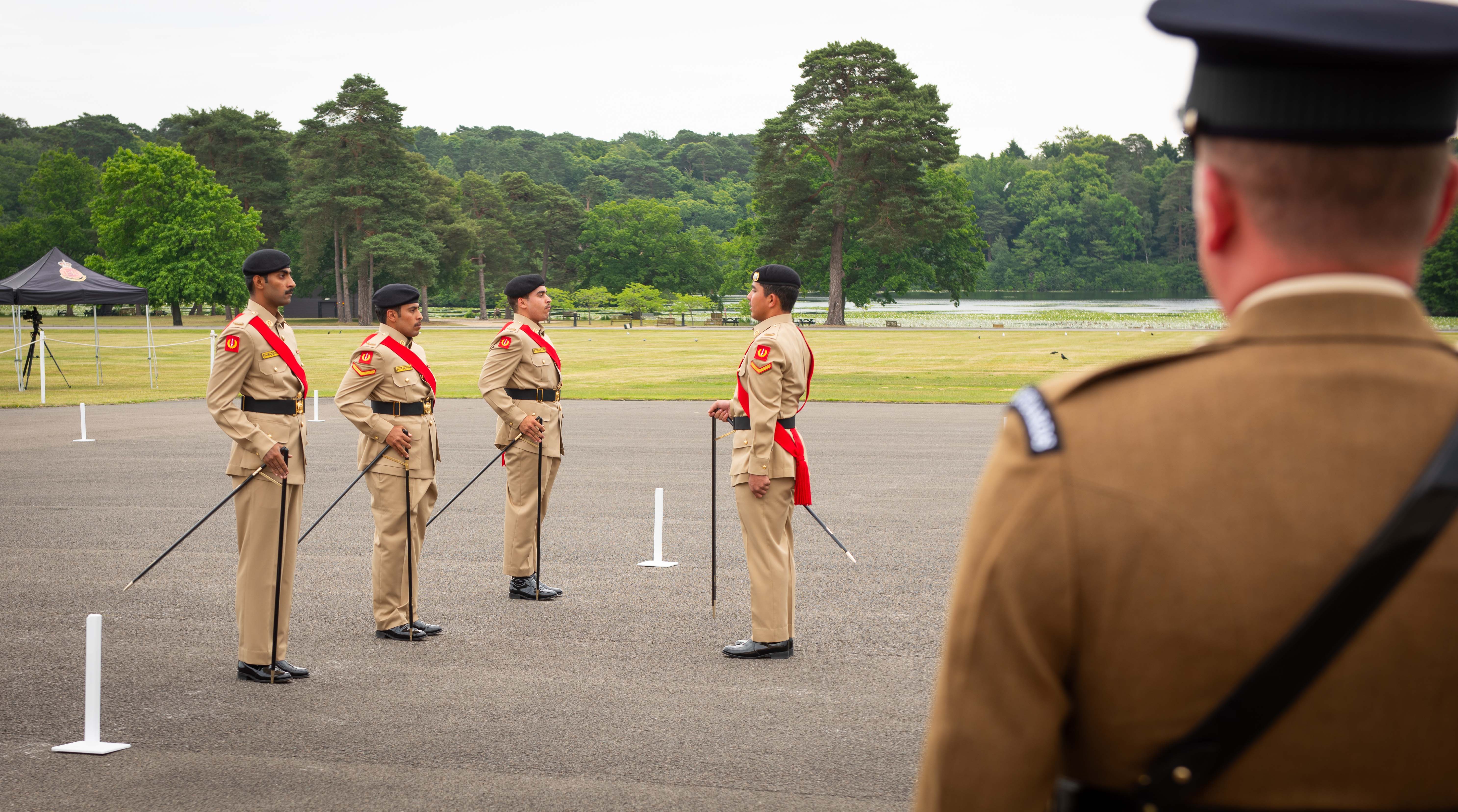 Four soldiers from the Bahrain Defence Force, dress uniforms are stood on a parade square carrying pace sticks. Looking at them in the foreground is a British Army soldier watching the team of four.