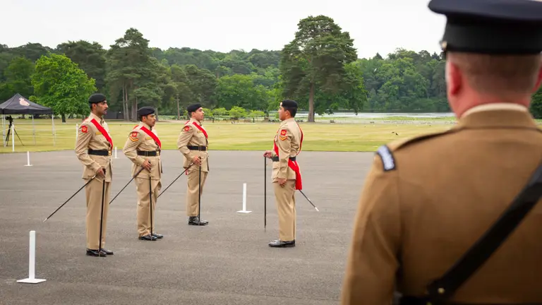 Four soldiers from the Bahrain Defence Force, dress uniforms are stood on a parade square carrying pace sticks. Looking at them in the foreground is a British Army soldier watching the team of four.